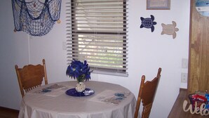 A dining area with a round table covered by a white tablecloth, two wooden chairs, a vase with blue flowers, and a window with blinds.