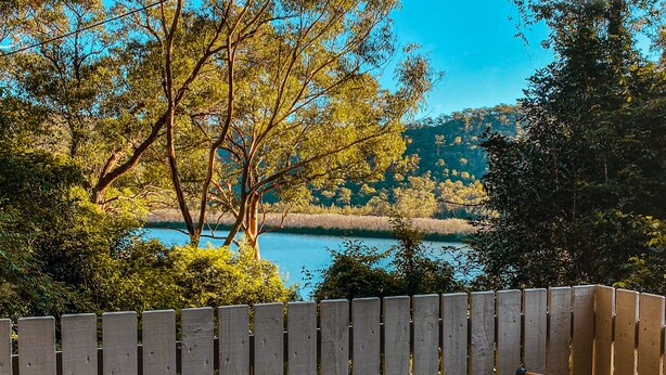 Un banco de madera con un libro y una taza, una cerca de madera, y una vista de un lago con árboles.