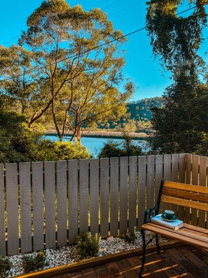 Un banco de madera con un libro y una taza, una cerca de madera, y una vista de un lago con árboles.