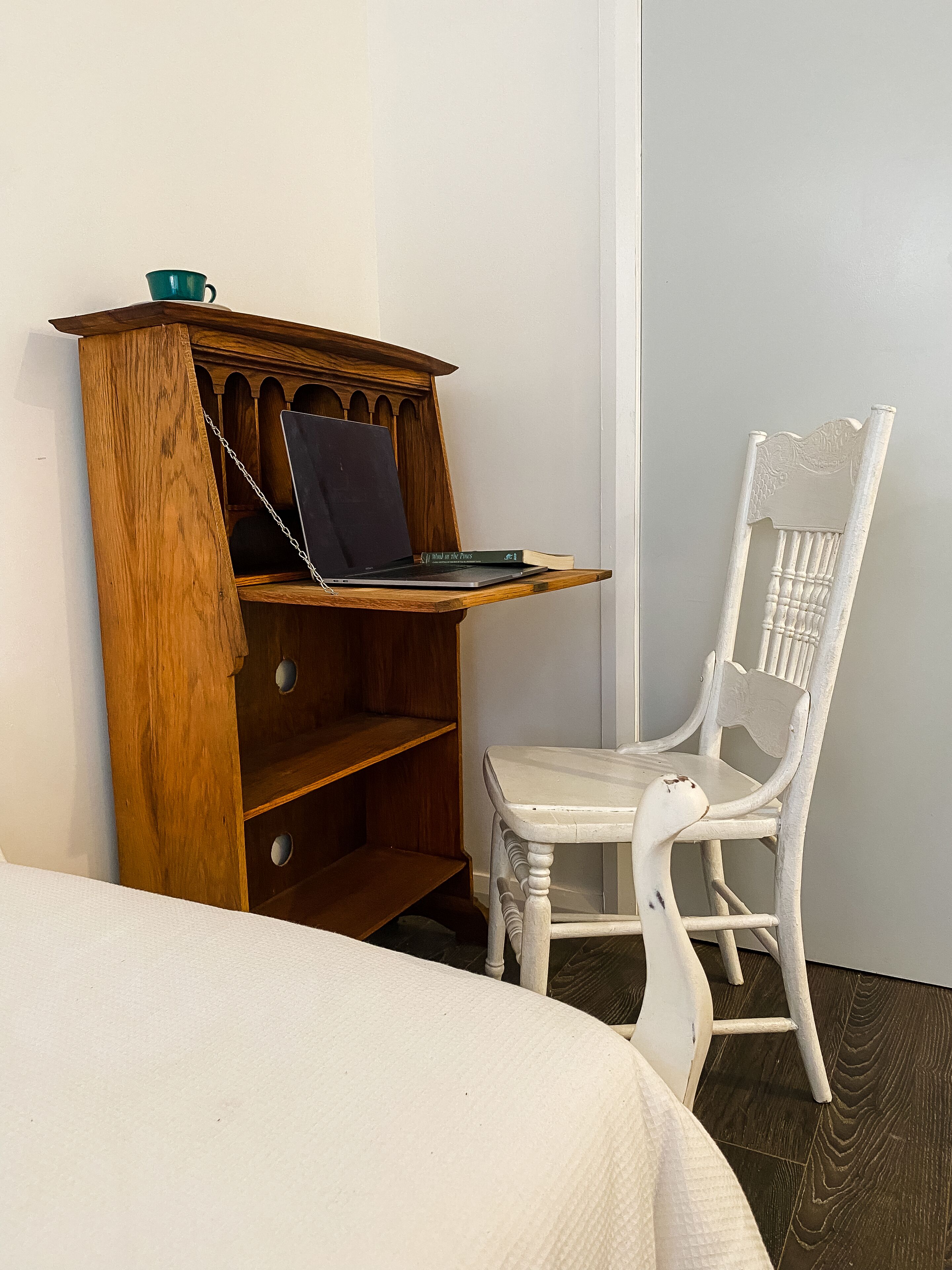 A wooden desk with a laptop and a book, a white chair, and a bed with a white cover.