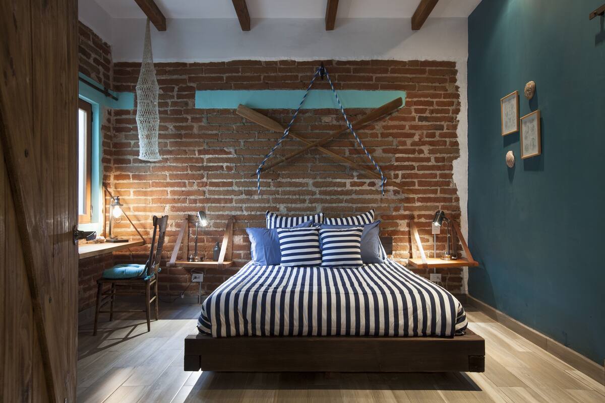 A bedroom with a brick wall, a wooden bed frame, a hanging light, and a desk with a chair.