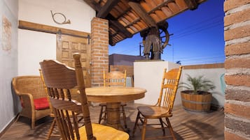 A dining area with a round wooden table, six wooden chairs, a wicker chair with a red cushion, a brick wall, a wooden statue, and a potted plant.