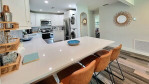 A modern kitchen with white cabinets, a central island with seating, and a dining area with a white table and orange chairs.