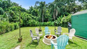 A backyard with a fire pit, white and blue Adirondack chairs, and a green shed.