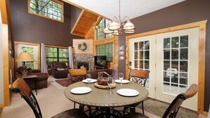 Un salón amplio con chimenea, un comedor con mesa y sillas redondas, y ventanas grandes que ofrecen vistas al exterior.