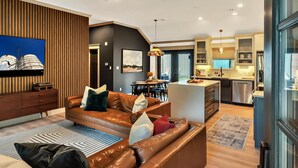 A modern living room with a leather sofa, a flat-screen TV, and a dining area with a kitchen in the background. - Lonetree Ranch an Outdoor Oasis w/ hot tub near Baylor/Magnolia (Waco)