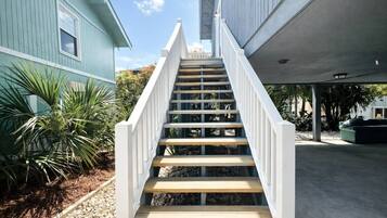 A staircase leading to a covered area with a concrete floor, surrounded by greenery and a building with a blue exterior.