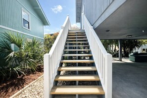 Un escalier mène à une zone abritée dotée d’un sol en béton, entourée de verdure et bordée d’un bâtiment à la façade bleue.