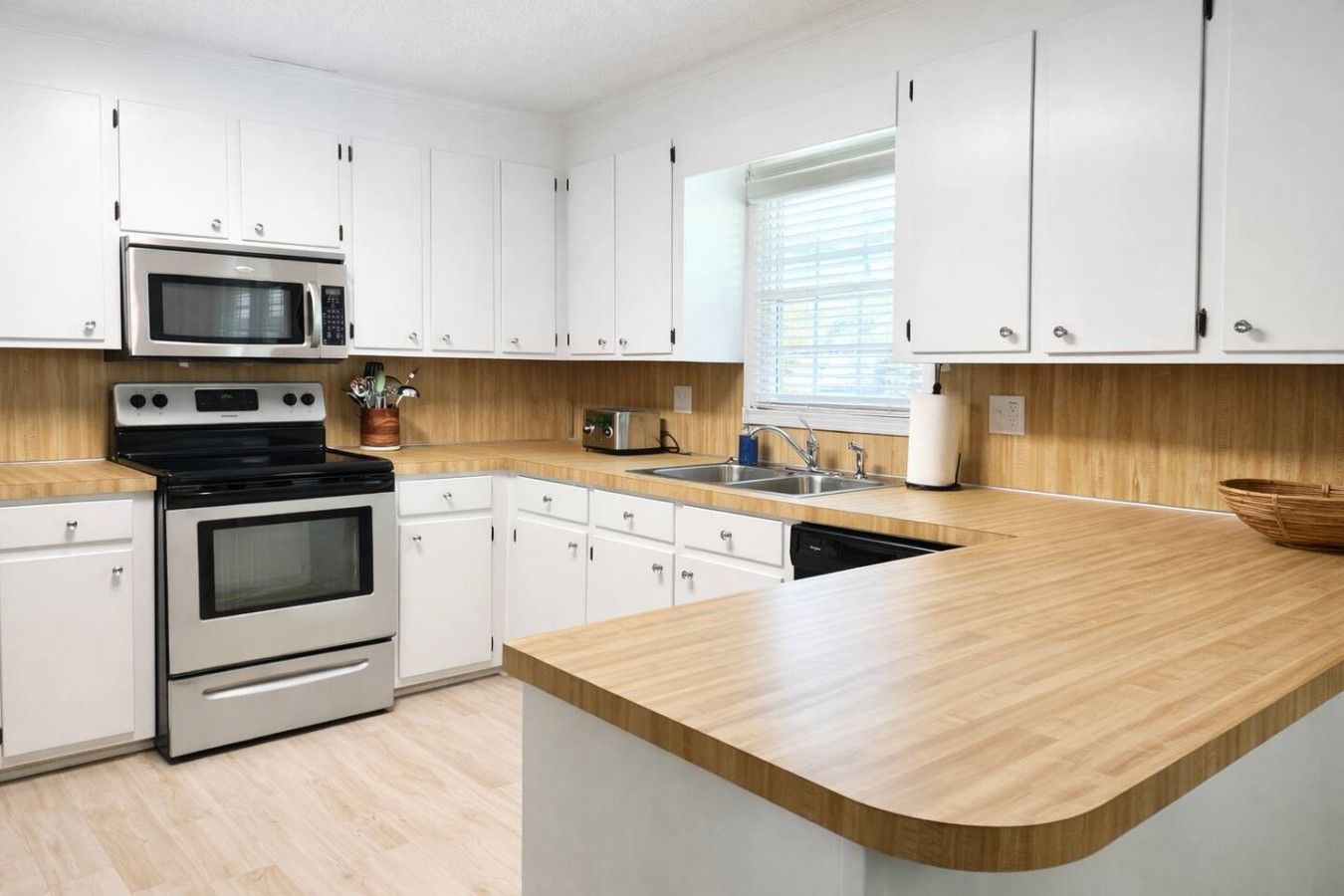 A modern kitchen with white cabinets, stainless steel appliances, a wooden countertop, and a window with blinds.