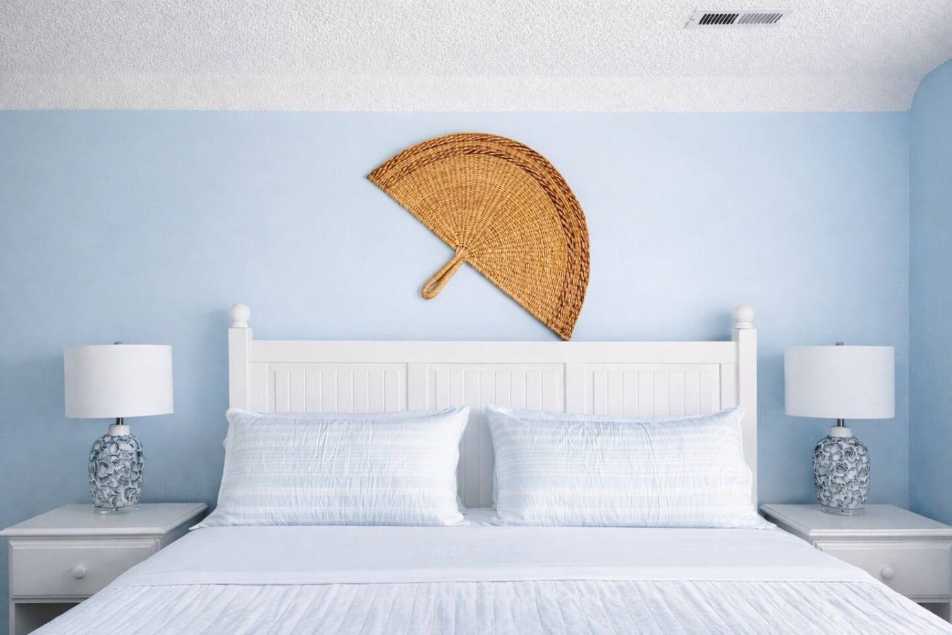 A bedroom with a white bed, two nightstands with lamps, and a woven fan on the wall.