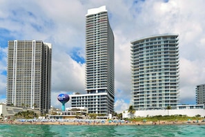 Un hotel frente a la playa con varios edificios altos, una playa con sombrillas y agua azul cristalina.