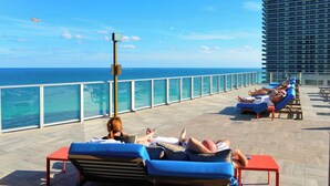 A rooftop lounge with sun loungers, a clear view of the ocean, and a tall building in the background. - Family 2BR | Beach Access | Sleeps 6 (Hollywood)