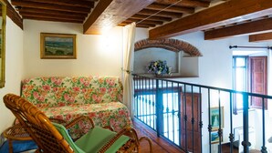 Une pièce avec un plafond en bois, un canapé à motifs floraux, des fauteuils en osier, un balcon avec une balustrade et une cheminée.