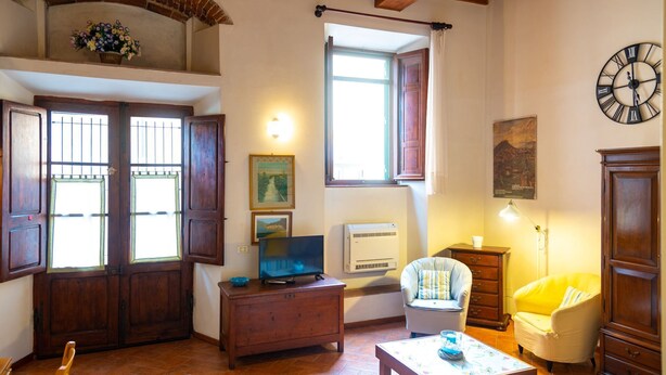 Un salon chaleureux avec une porte en bois, un téléviseur posé sur un meuble en bois, un fauteuil blanc, un fauteuil jaune, une horloge au mur et une fenêtre avec des rideaux.