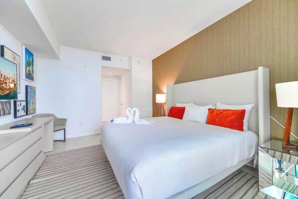 A hotel room with a large bed, a desk, a chair, a lamp, and artwork on the wall.