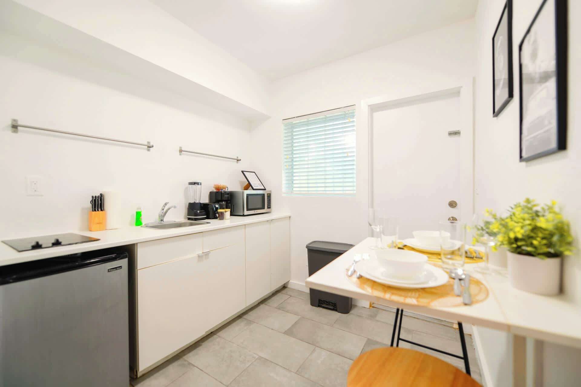 A compact kitchen with a dishwasher, microwave, and coffee maker. There is a small trash bin and a window with blinds.