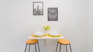 A dining area with a white table set for two, featuring yellow placemats, white plates, and a vase with yellow flowers. Two wooden stools with black metal legs are positioned at each end of the table. - Miami Studio | Minutes from Design District (Miami)