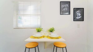 A dining area with a white table set for two, two wooden stools, a window with blinds, and two framed pictures on the wall. - Stay Minutes from Miami Hotspots (Miami)