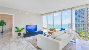 A modern living room with a large TV, comfortable seating, and a view of the ocean and buildings. - 2BR Bay-View Brickell | Steps to City Centre (Miami)