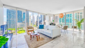 A spacious living room with a white sofa, a coffee table, and a dining area with chairs. Large windows offer a view of tall buildings. - 2BR Bay-View Brickell | Steps to City Centre (Miami)