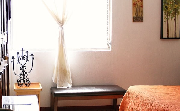 A room with a bed, a bench, a small table with a candelabra, and a window with sheer curtains.