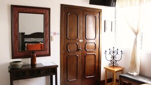 Una habitación con puerta de madera, un espejo, una mesa pequeña con un candelabro y una cama con un cojín rojo.