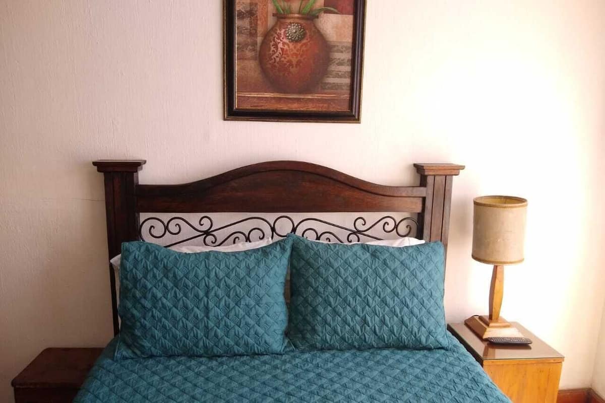 Un lit avec une tête de lit en bois et deux coussins de couleur turquoise, une table de chevet avec une lampe et un tableau encadré accroché au mur.