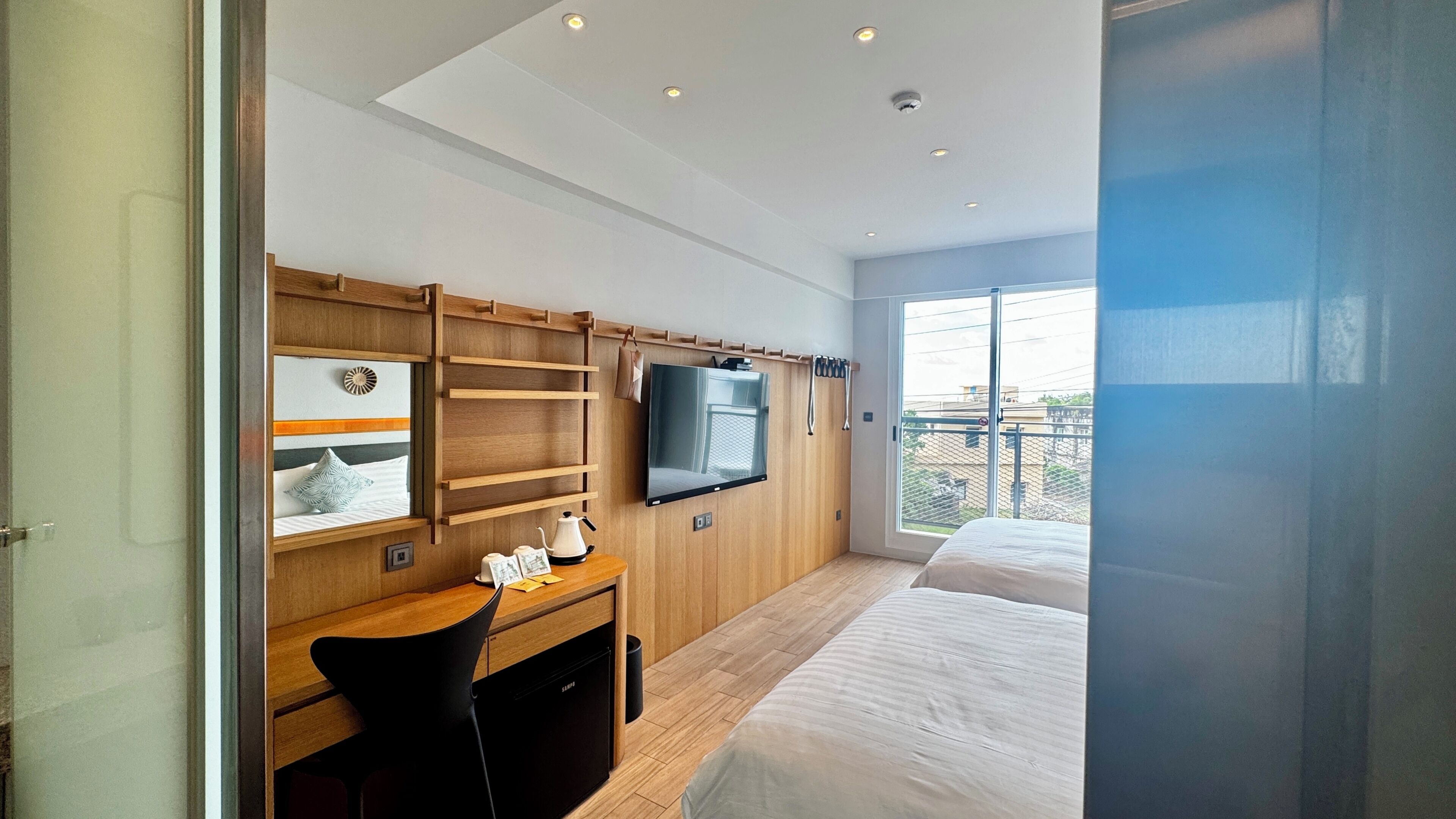 A modern hotel room with a bed, a desk with a chair, a television, and a large window.