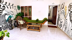 A room with a green cushioned bench, a wooden chair, a small table, and a wall with black and white floral patterns. - Aryavilla Heritage (Varkala)