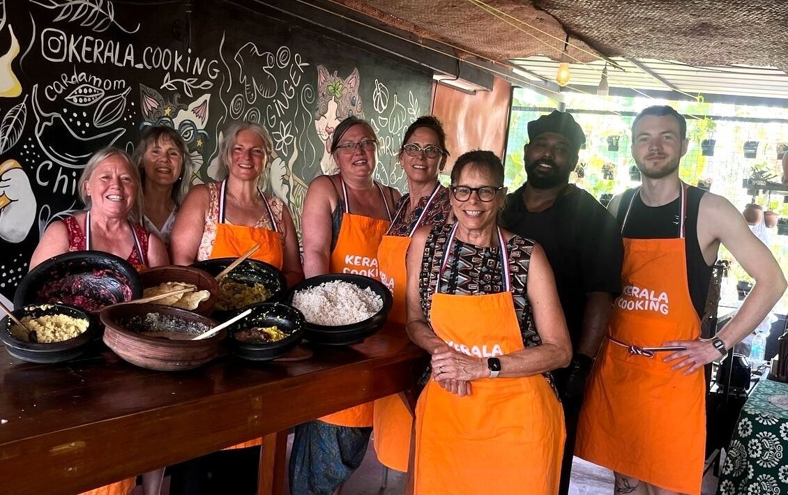 En gruppe mennesker iført orange forklæder med teksten 'KERALA COOKING', står foran en tavle med tegninger og ord relateret til mad.