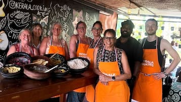 En gruppe mennesker iført orange forklæder med teksten 'KERALA COOKING', står foran en tavle med tegninger og ord relateret til mad.