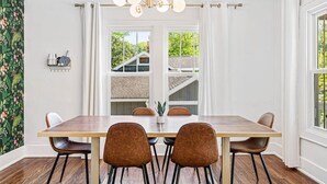A dining room with a wooden table, six chairs, a chandelier, and a view of a house through a glass door.