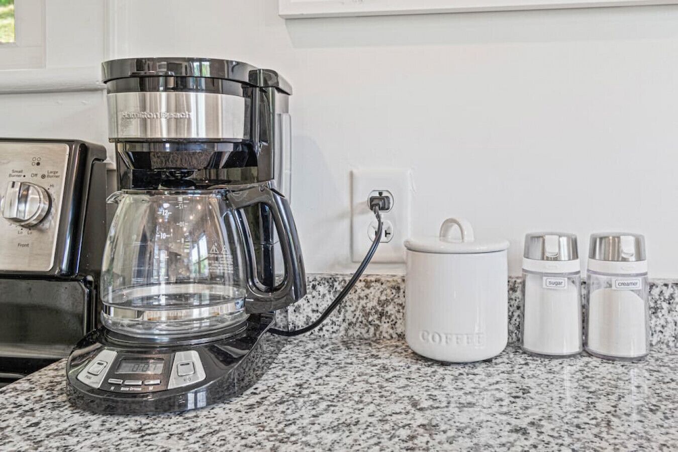 Een koffiezetapparaat op een granieten aanrecht, met een koffiekan, een koffiezetapparaat en twee potjes met 'suiker' en 'room'.