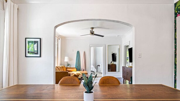 A spacious room with a wooden dining table, a ceiling fan, and a view into a living area with a sofa and a television.