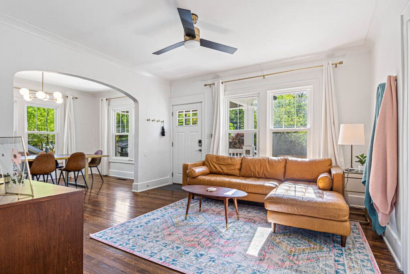 A spacious living room with a brown leather sofa, a coffee table, and a patterned rug. There is a dining area with a table and chairs, and a ceiling fan.