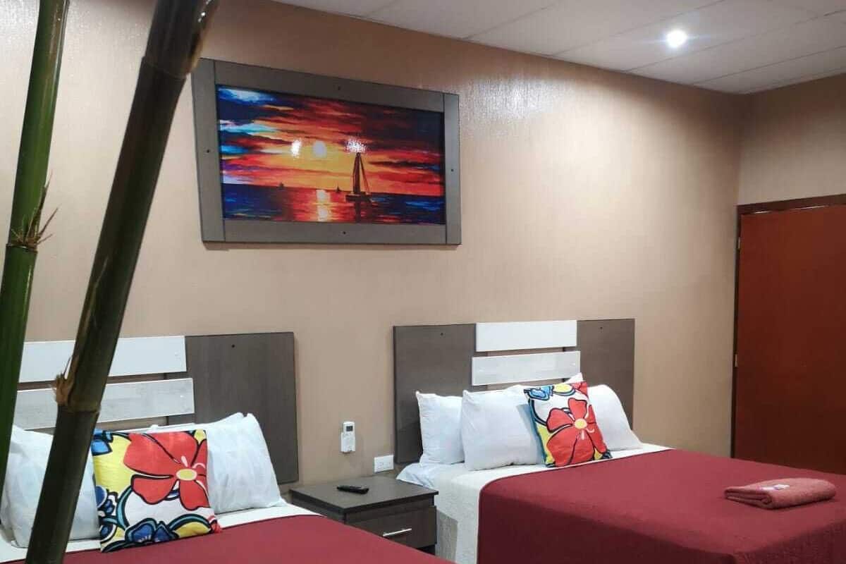 A hotel room with two beds, each with a red bedspread and a colorful pillow. There is a painting of a sunset on the wall, a bamboo plant in the corner, and a tiled floor.