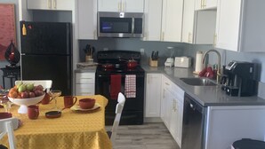 Une cuisine moderne avec des meubles blancs, un réfrigérateur en acier inoxydable, un four à micro-ondes et une cuisinière. On y trouve une table à manger recouverte d’une nappe jaune et un saladier rempli de fruits.