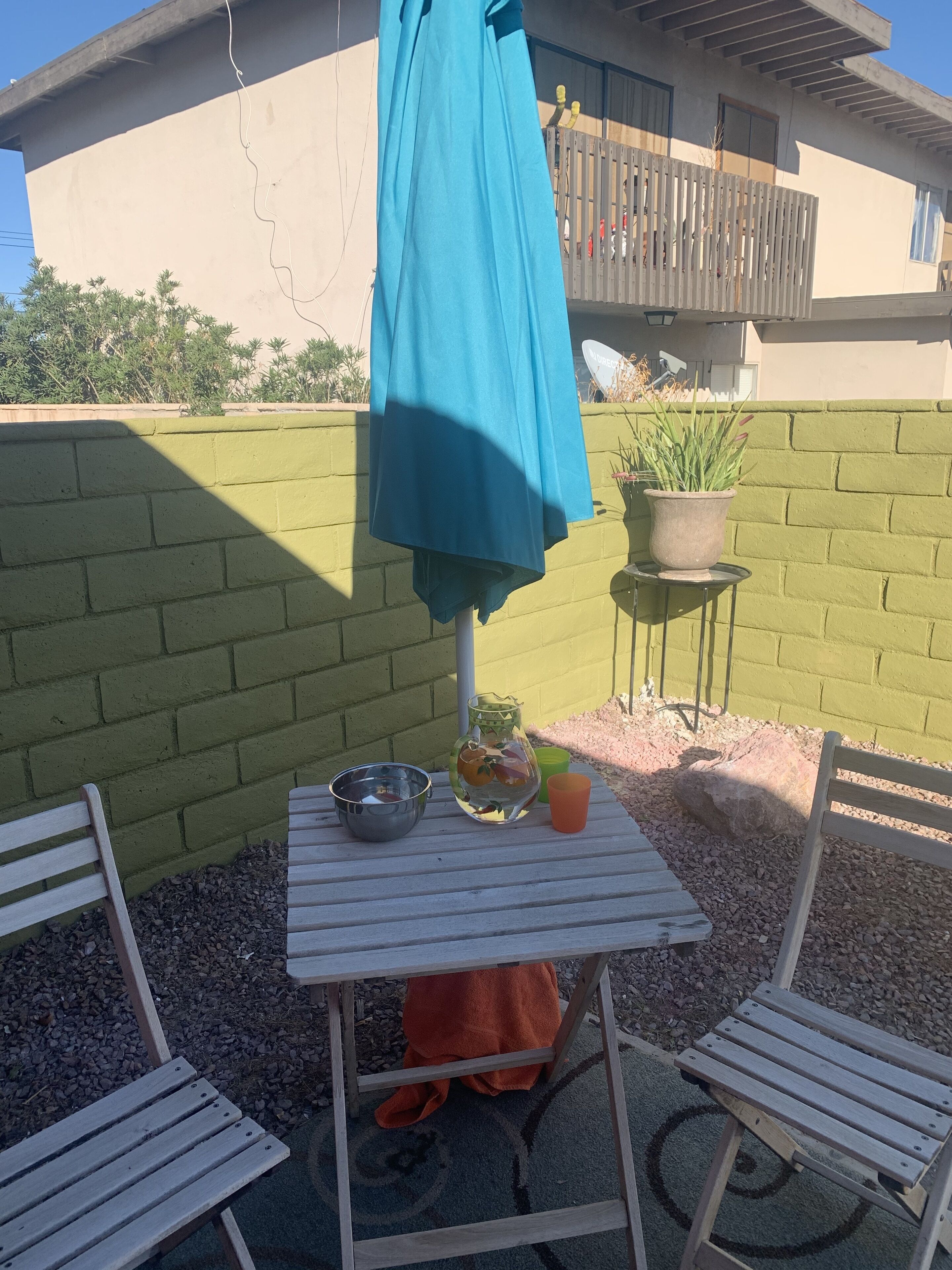 Un pequeño patio con una mesa, sillas, un paraguas azul, una planta en maceta y un muro de ladrillo.