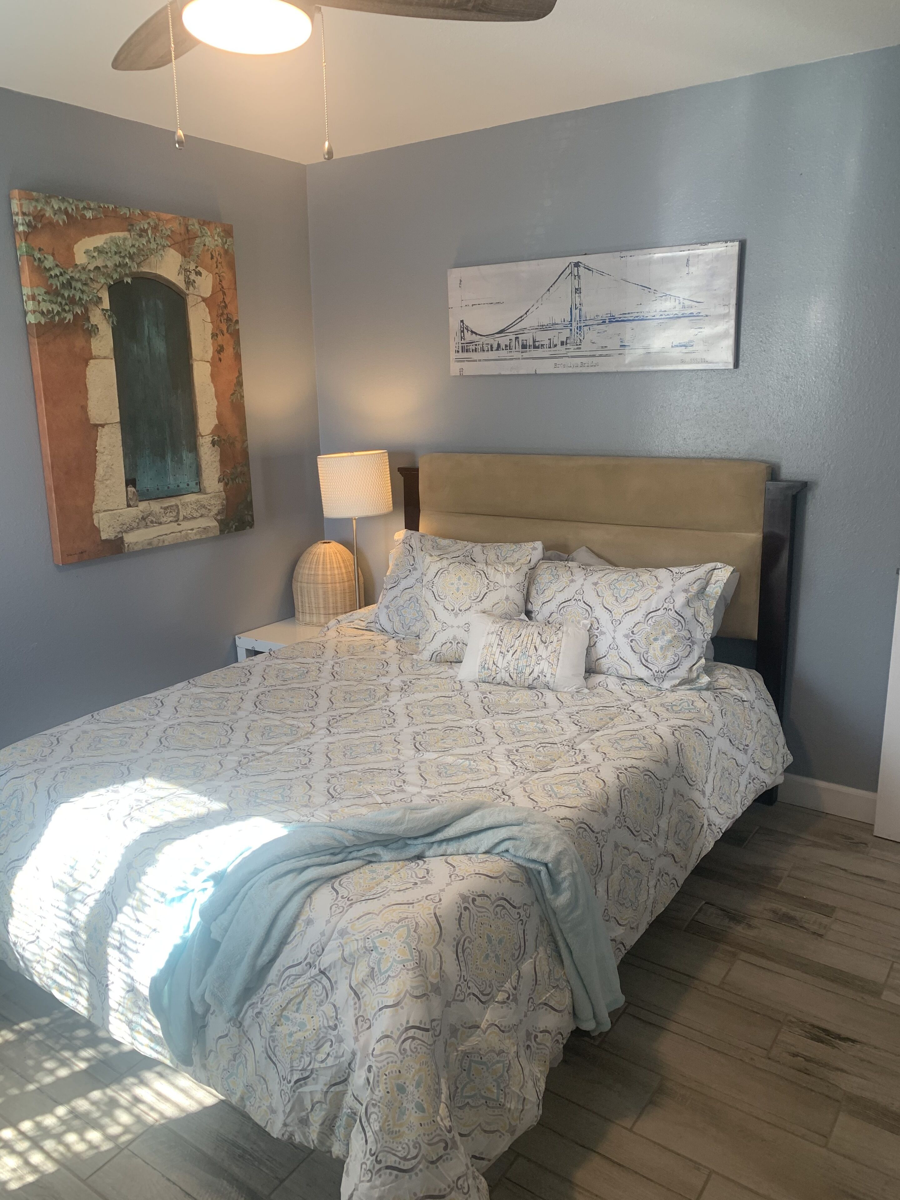 A bedroom with a bed, a nightstand, a lamp, a ceiling fan, and artwork on the walls.