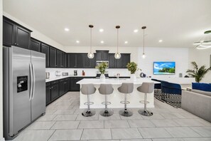 A modern kitchen with dark cabinets, a stainless steel refrigerator, a central island with bar stools, and a flat-screen TV on the wall. - Secluded 10 Bedroom Villa With Ocean Views 6282 (Davenport)