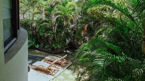 Un espace extérieur abrité, doté d’un toit de chaume, entouré d’une végétation tropicale luxuriante et agrémenté d’une terrasse en bois avec des balustrades blanches.