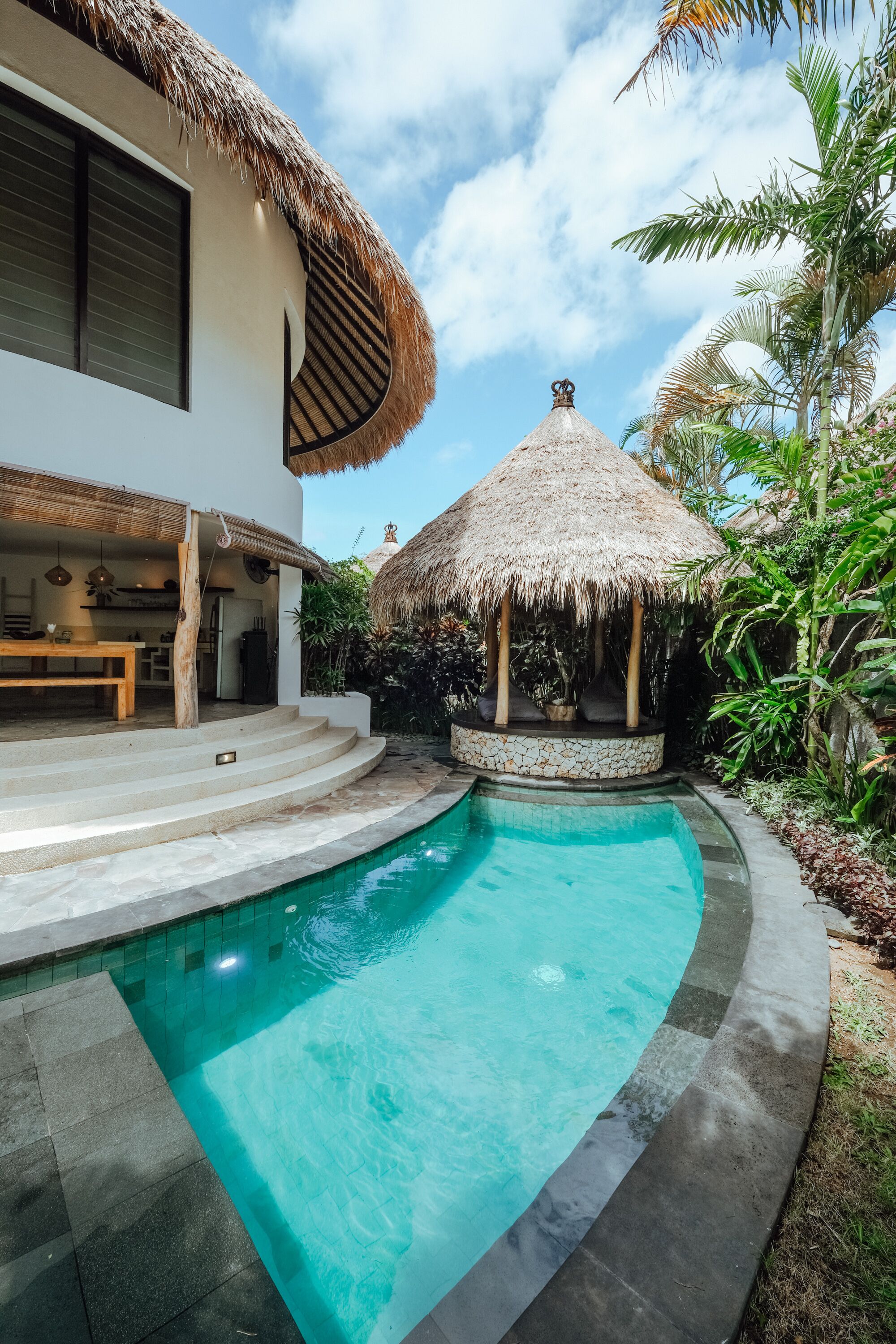 A swimming pool with a curved edge, a thatched-roof structure, and a modern house with a large window.