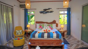 A hotel room with a large bed, a wooden headboard, a yellow armchair, a wooden bench, and a dining table. - Milles Montagnes Residence (Mahé Island)