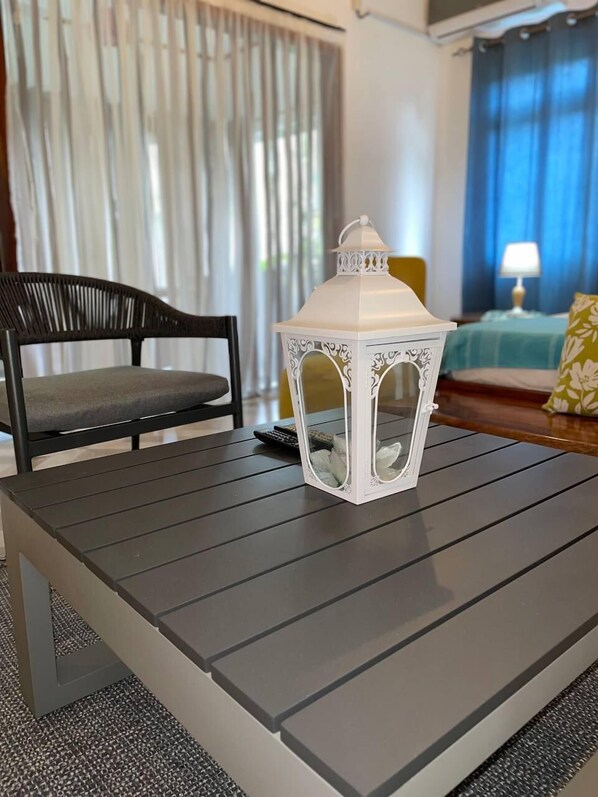 A modern living room with a dark wooden coffee table, a white decorative lantern, a black chair, and a bed with blue curtains in the background. - Milles Montagnes Residence (Mahé Island)