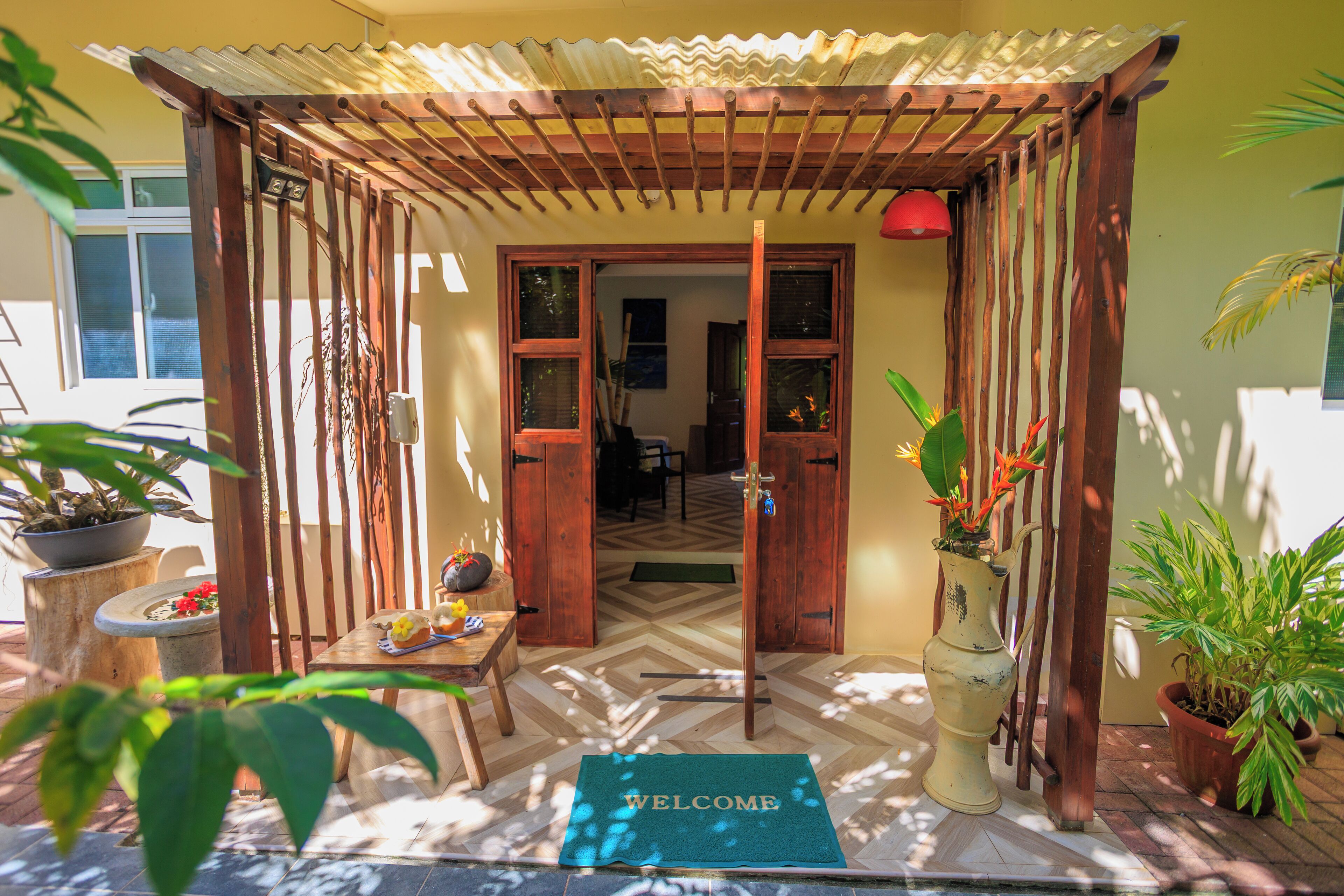 A wooden pergola entrance with a welcome mat, potted plants, and a tiled floor.