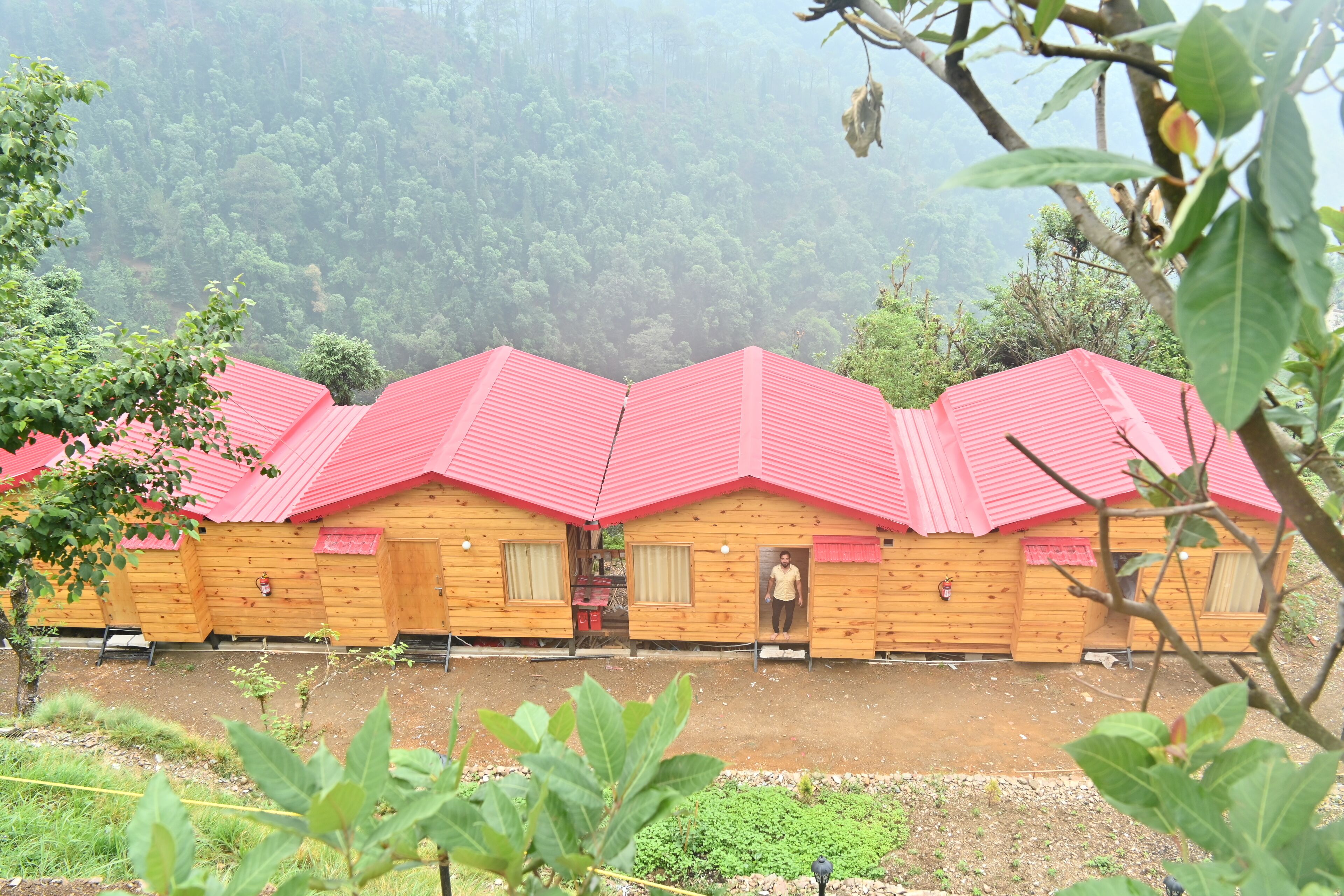 A row of wooden cabins with red roofs, set against a forest backdrop.