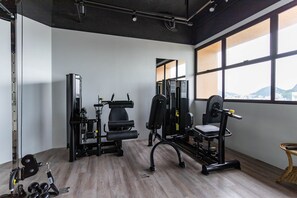 Un gimnasio con diversos equipos de ejercicio, grandes ventanales y vistas a un paisaje urbano.