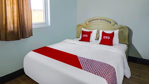 A neatly made bed with a white and red bedspread, two pillows with 'OYO' branding, a window with brown curtains, and a light blue wall. - Hotel O Guesthouse Rantang (Medan)
