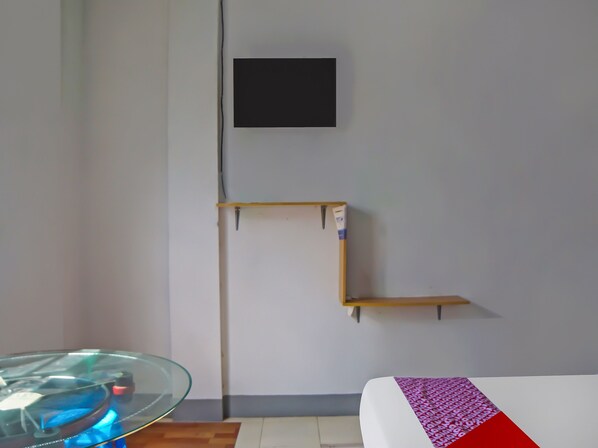 A room with a bed, a glass table with a blue object on it, and a wall-mounted shelf with a box.
