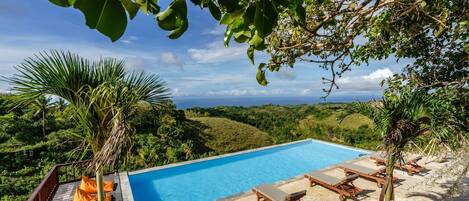 A clear blue pool with lounge chairs and a wooden deck, surrounded by lush greenery and hills.